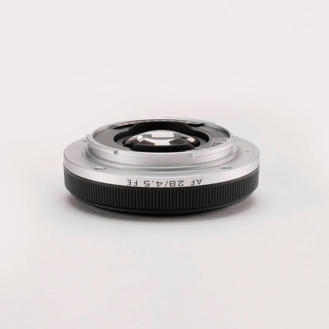 A silver and black Viltrox 28mm f/4.5 FE wide-angle lens for Sony full-frame cameras lies flat on a white surface, showcasing its compact build, ribbed black grip, visible glass elements, and minimalistic background.