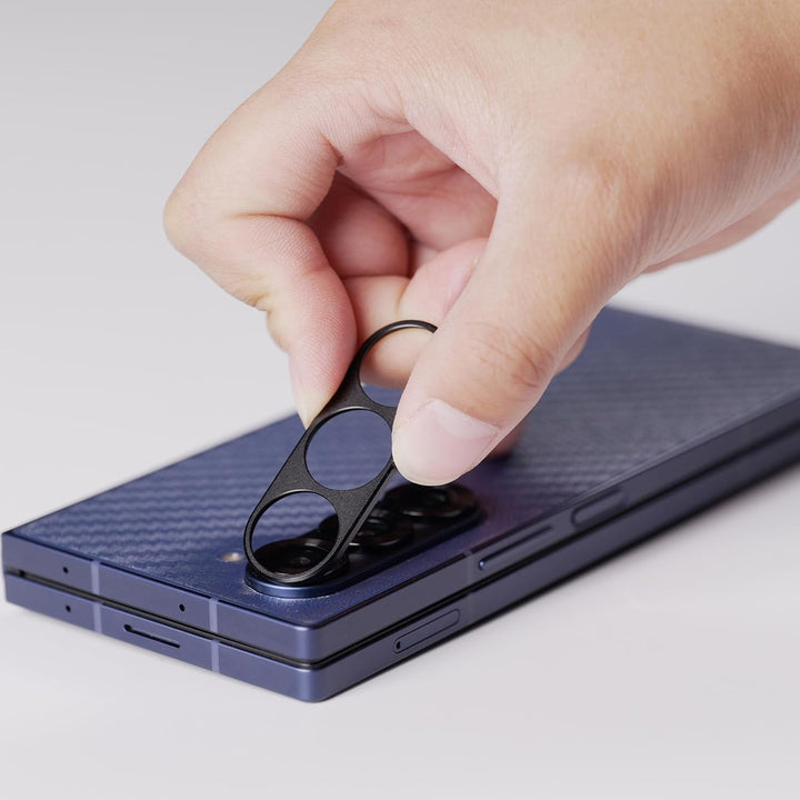 A hand places a MobileTechnica Samsung Galaxy Z Fold 7 camera lens protector onto the blue, folded phone’s module, showing the 9H tempered glass cutouts and alignment on a light background.