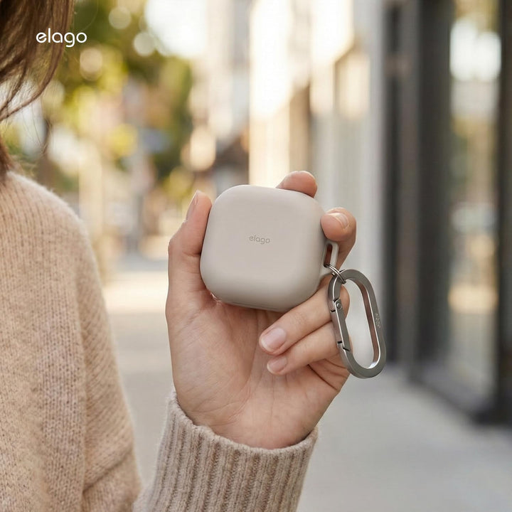 A person in a beige sweater holds the MobileTechnica Samsung Galaxy Buds 4 Pro & Galaxy Buds 4 Case (Silicone, Full Protection, Wireless Charging, Stone) with a metal carabiner on a city sidewalk amid softly blurred buildings and greenery.