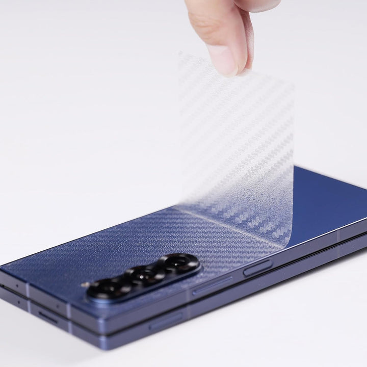 A hand peels a MobileTechnica Samsung Galaxy Z Fold 7 tempered glass screen protector with camera lens cover from the back of a folded, blue phone with triple cameras, showing the buttons and hinge on a white surface.