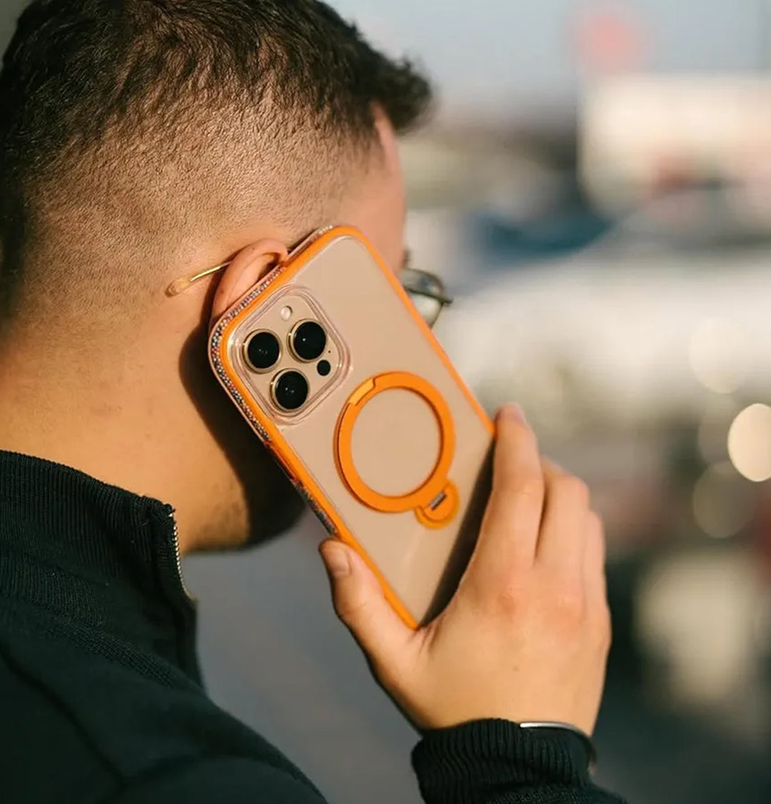 A short-haired individual holds the Raptic AirJoy Beads with Magsafe for iPhone 16 Pro Max to their ear, flaunting its clear case with air bumpers and an orange ring. Sporting clear-framed glasses and a dark jacket against a sunny blurred backdrop, they exude a modern vibe.