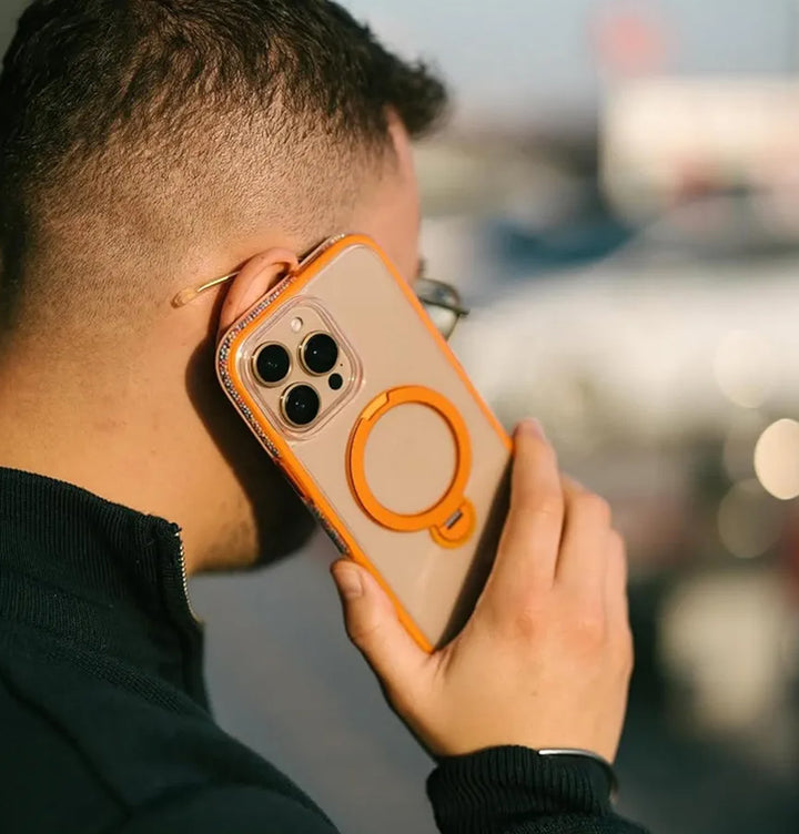 A person stands outdoors, wearing glasses and a dark jacket, holding an iPhone 16 Pro with Raptic's AirJoy Beads case in orange. The eye-catching bead design features a circular 360 kickstand on the back, and the phone's multiple camera lenses are visible against a blurred background.