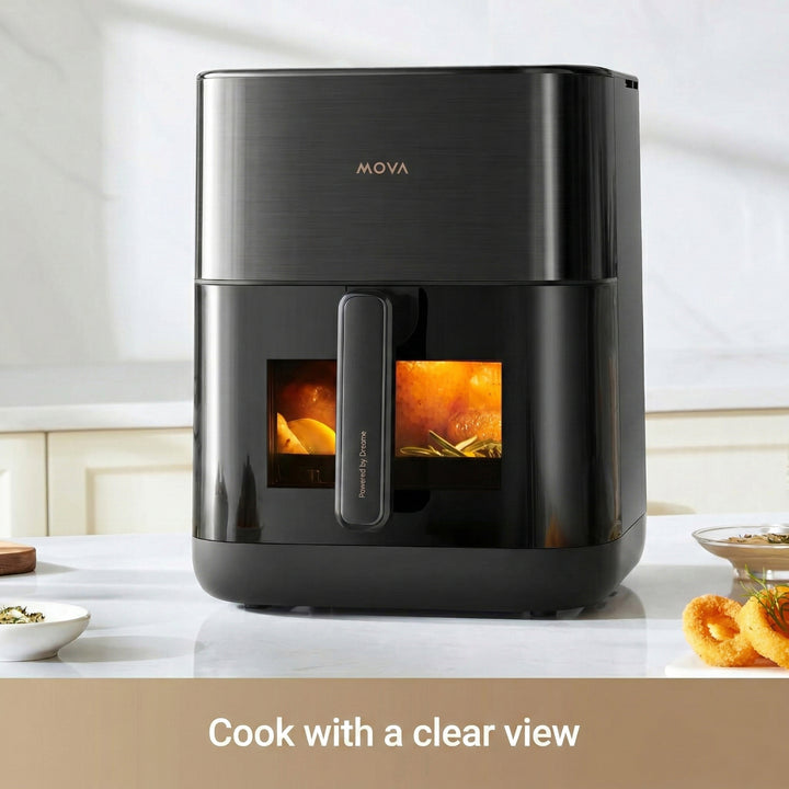A sleek black MobileTechnica MOVA FD10s Pro Air Fryer with Dual HeatSync System sits on a kitchen counter. Two clear windows show food crisping inside, with gold accents on the handle and logo. Text reads, “Cook with a clear view.”.