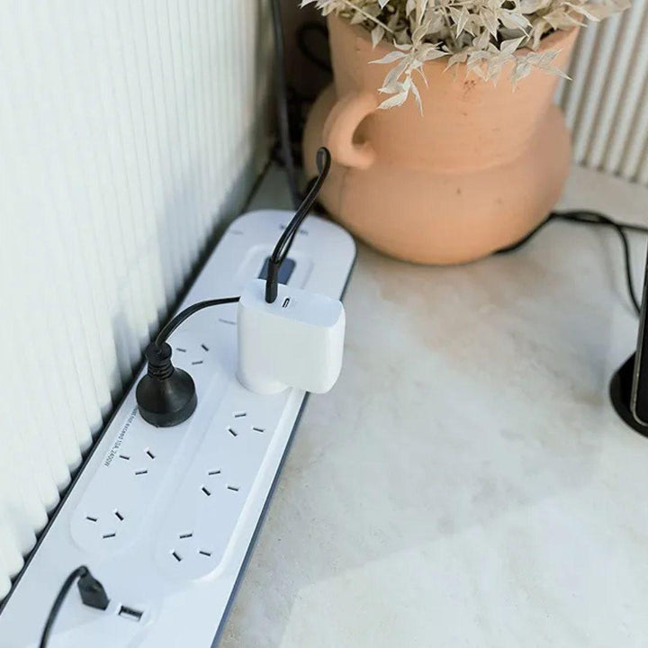 A MobileTechnica Surge Plus USB power strip with surge protection and multiple outlets, including USB ports, sits on a light floor by a beige wall. Two plugs—one black, one white with a large adapter—are connected nearby a peach pot with dried leaves.