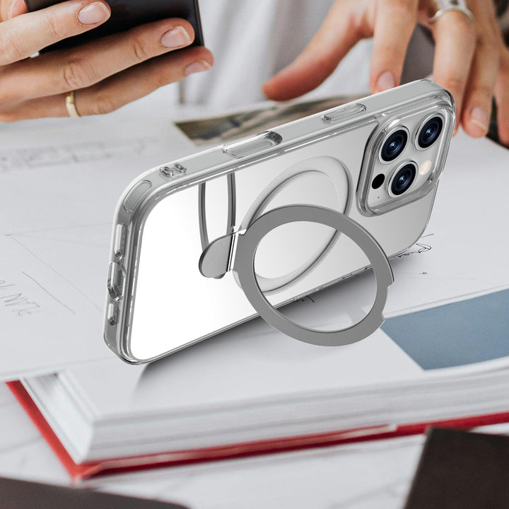 The Raptic Air Stand Mirror Msafe iPhone 16, a clear 3-in-1 phone case with a built-in ring holder, is propped up on its side. The ring holder, attached to the back of the case, doubles as a kickstand. In the background, a person's hands are busy using another smartphone amidst scattered papers and notebooks on the table.