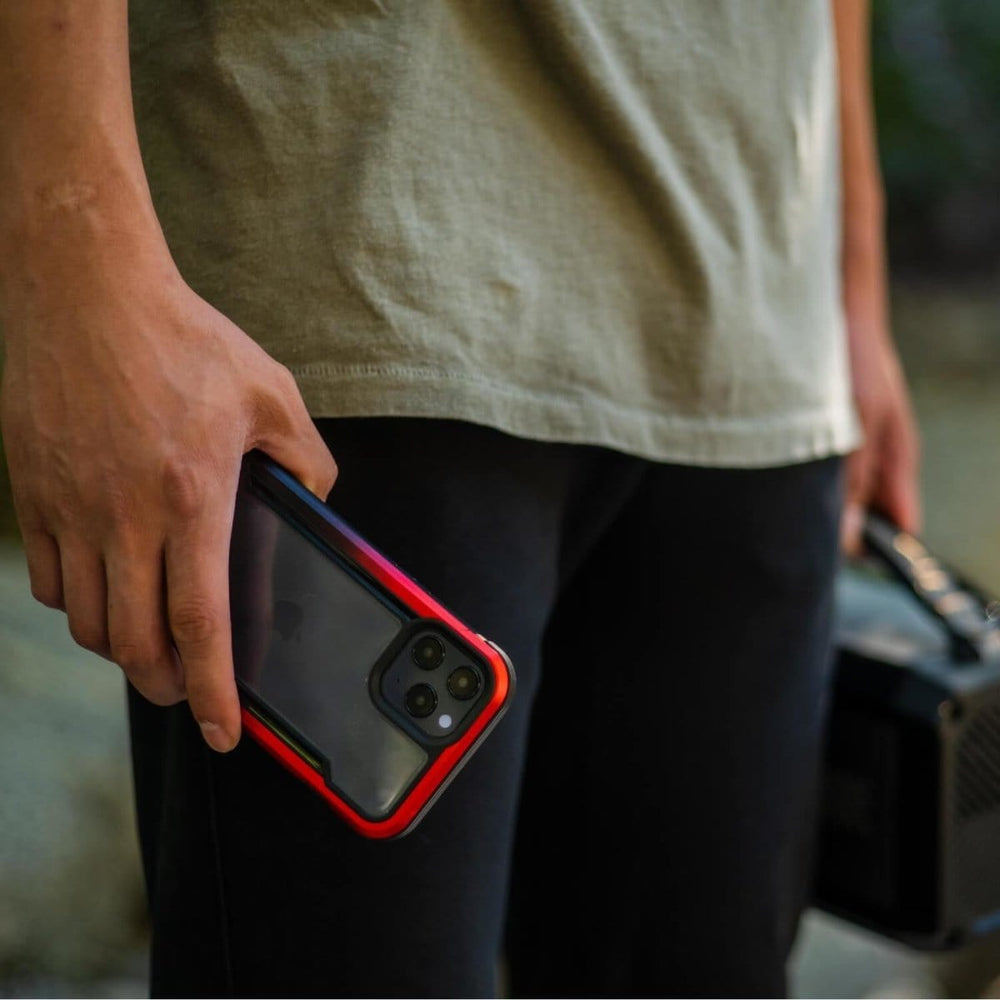 A person wearing a beige shirt and black pants holds a smartphone with an iPhone 12 Pro Max Case - Raptic Shield red and black case in one hand and a portable speaker in the other.