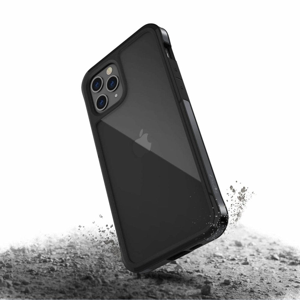 A black Raptic Edge iPhone 12 Pro Bumper Case with a rugged, durable design and enhanced corners is shown. The phone inside, boasting a transparent back cover and machined aluminum frame, is depicted falling onto a rough, dusty surface, emphasizing its protective features.
