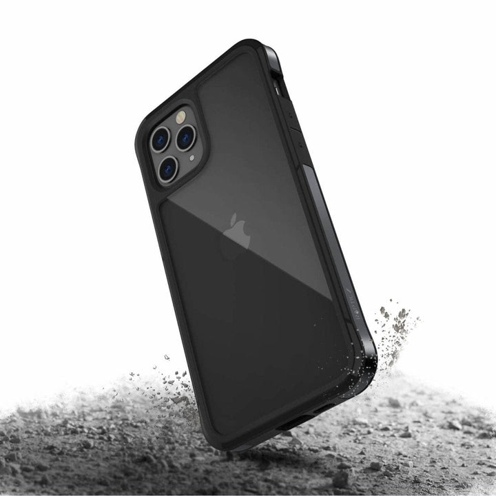 A black Raptic Edge iPhone 12 Pro Bumper Case with a rugged, durable design and enhanced corners is shown. The phone inside, boasting a transparent back cover and machined aluminum frame, is depicted falling onto a rough, dusty surface, emphasizing its protective features.