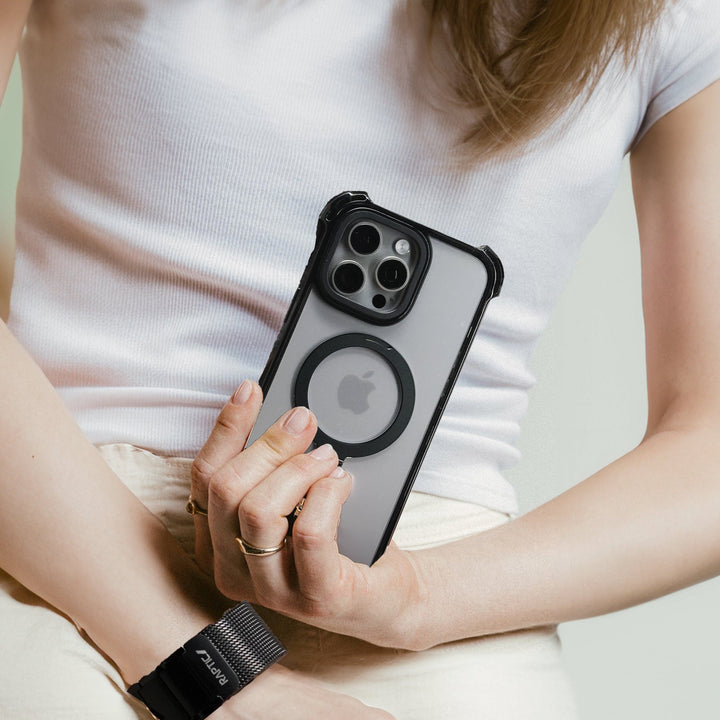 A person wearing a white sleeveless top holds a Raptic Dual Impact Kickstand Case for iPhone 16 Pro with ultimate drop protection and a black bumper featuring a circular magnetic ring on the back, making it MagSafe-compatible. The individual wears a black smartwatch and a gold ring on their right hand, while their left arm is wrapped around their torso against a plain, light background.