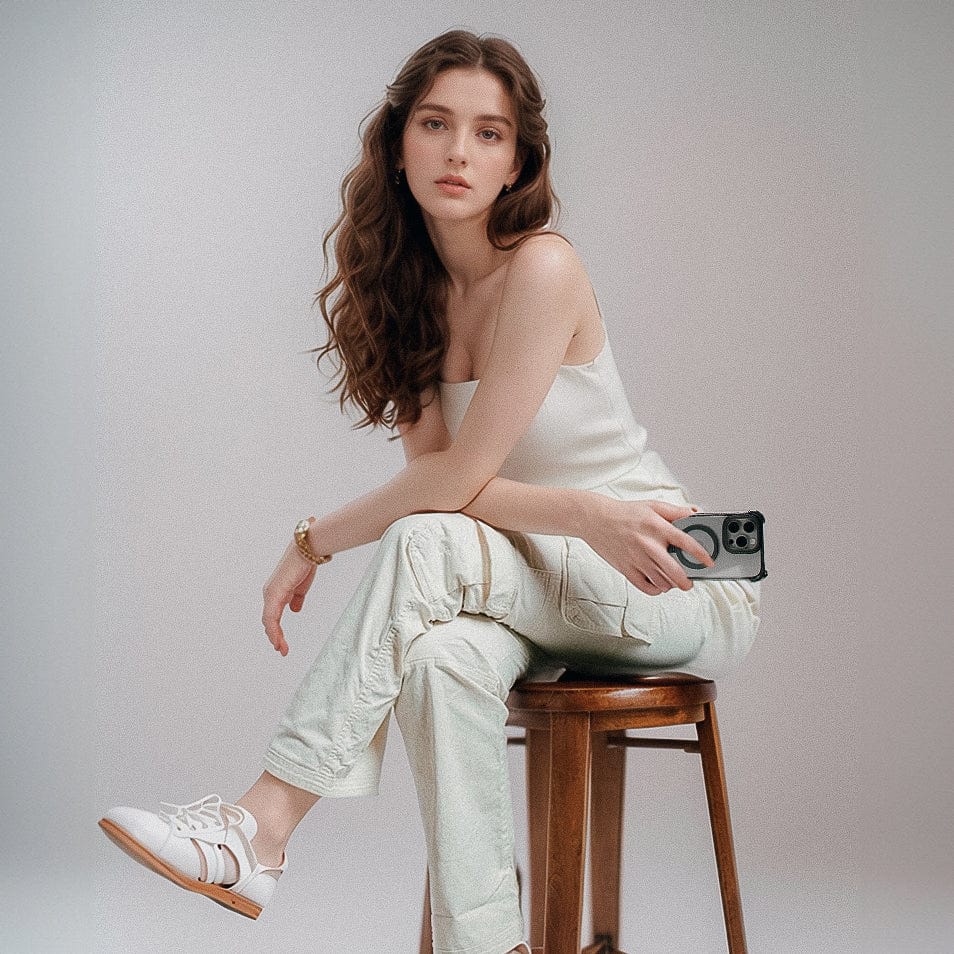 A young woman with long wavy hair sits on a wooden stool against a plain background. She wears a white tank top, light-colored cargo pants, and white sneakers. Holding a Raptic Dual Impact Kickstand Case for iPhone 16 Pro in her left hand, her elbow rests on her knee as she gazes towards the camera.