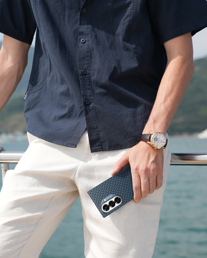 A person in a navy short-sleeve shirt and beige pants holds a RAPTIC Samsung Galaxy Fold 7 Skin Case - Profound Navy by the railing, wearing a watch with a white face and brown strap. Water and blurred hills appear in the background.