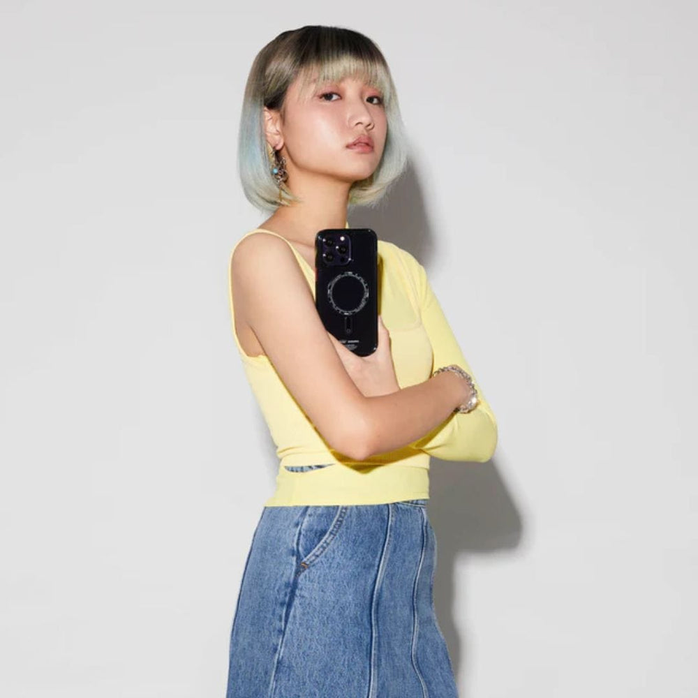 A person with a bob hairstyle dyed light blue and grey, wearing a yellow top and blue jeans, stands against a plain white background, holding an iPhone 14 Pro Max SAIDO Case - Skinarma from Skinarma with 360 full protection and scratch resistance against their chest.