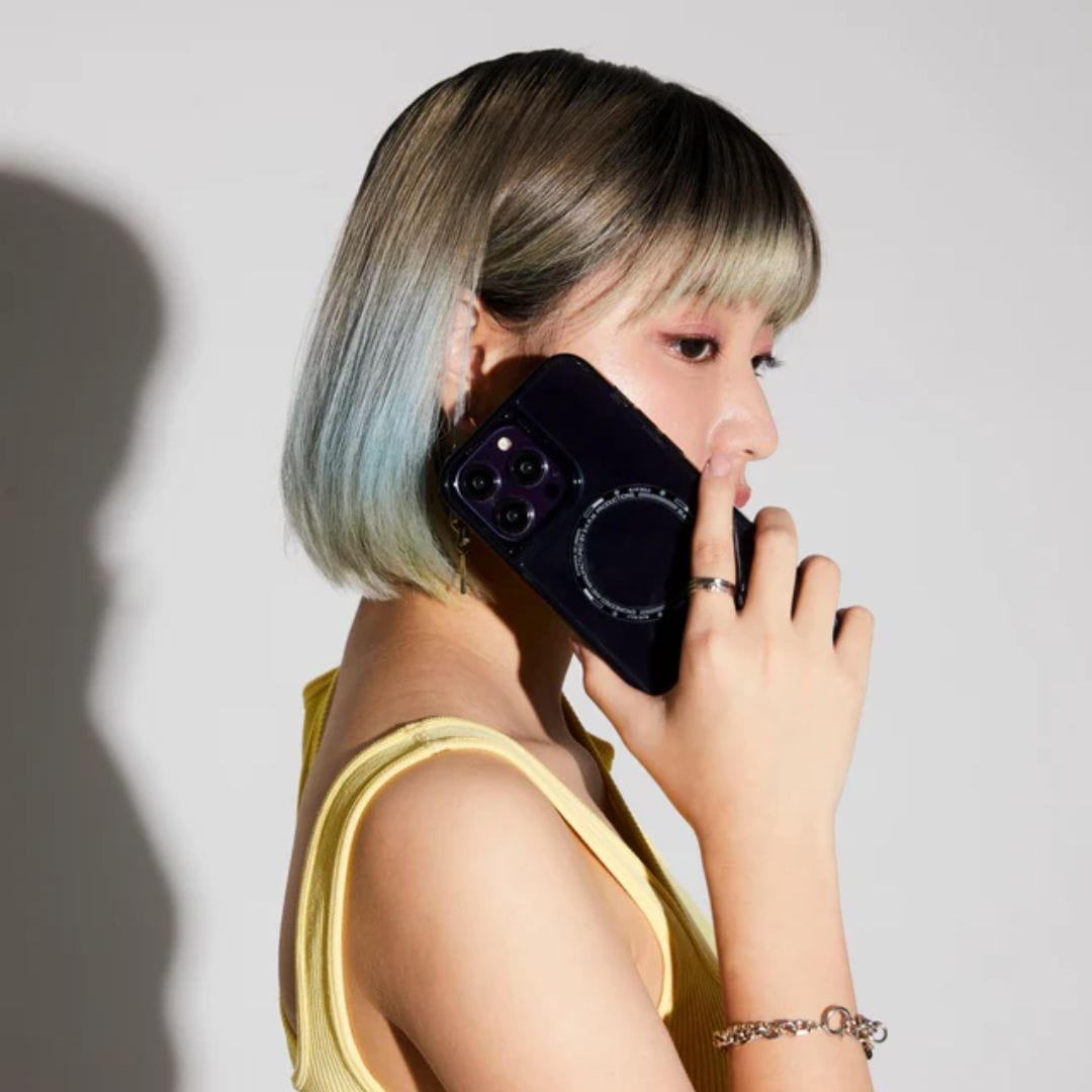A person with short hair holding a smartphone to their ear while wearing a sleeveless yellow top, seen from a side view. The phone, an iPhone 14 Pro Max SAIDO Case - Skinarma by Skinarma, featuring 360 full protection, seems perfect for this busy moment.
