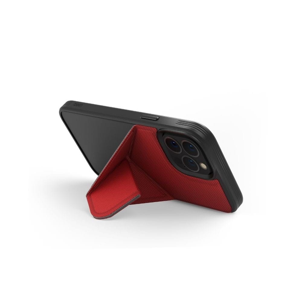 The iPhone 13 is propped up horizontally using a red UNIQ Transforma MagSafe Folding Case attached to its back. The case, shaped like a triangular prism, provides stable support for hands-free use. The phone's rear camera lenses feature a triple camera setup. The white background accentuates the phone and stand while ensuring drop protection.