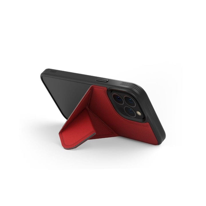 The iPhone 13 is propped up horizontally using a red UNIQ Transforma MagSafe Folding Case attached to its back. The case, shaped like a triangular prism, provides stable support for hands-free use. The phone's rear camera lenses feature a triple camera setup. The white background accentuates the phone and stand while ensuring drop protection.